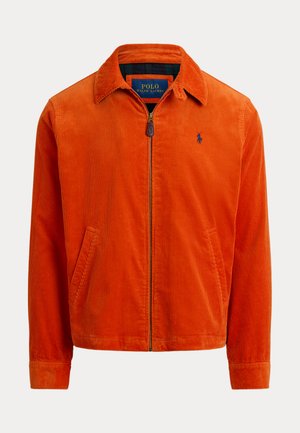 Jeune homme portant une casquette violette, une veste orange, un gilet en tricot à motifs, une chemise rayée, un pantalon cargo olive et des bottes marron, debout devant un fond beige.