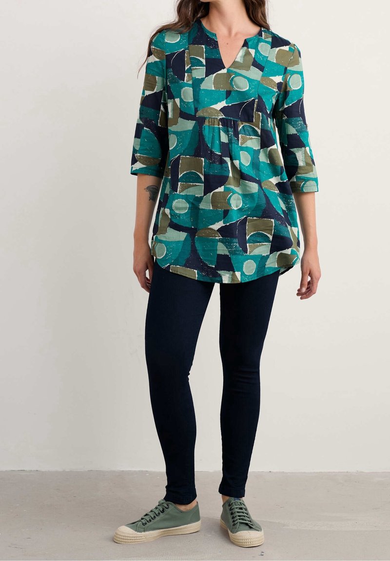 Femme portant une tunique à motif abstrait bleu sarcelle, marine et olive avec manches trois-quarts, leggings foncés et baskets vertes, debout à l’intérieur.