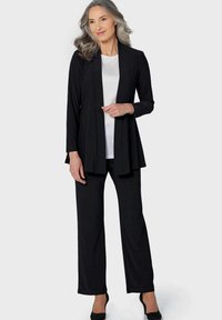 Schwarzes Outfit mit einer langen offenen Strickjacke, einem weißen Oberteil mit dezenten Verzierungen und geraden Hosen, abgerundet mit schwarzen Absätzen.