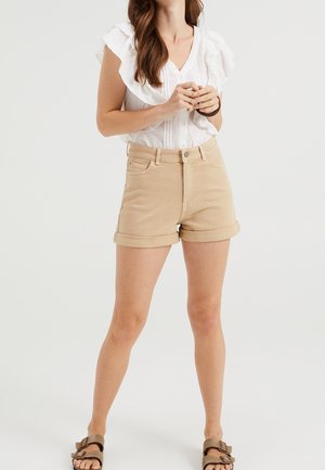 Kvinde iført beige opkrogede shorts, hvidt flæset kortærmet bluse og brune sandaler med stropper, stående med hænderne foldede.