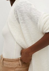 Valkoinen teksturoitu neuletakki, joka on heitetty kevyen beigehtävän istuvan pitkähihaisen paidan päälle, yhdistettynä vaaleanruskeisiin housuihin. Käsi säätää vyötärönauhaa.