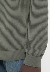 Vihreä ribattu neule, jossa on teksturoitu kangas. Hihansuut ja helma ovat istuvat, yhdistettyinä vaaleansinisiin farkkuihin. Ei huomattavia kuvioita tai varusteita.