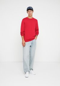 Pull en tricot rouge à manches longues, associé à un jean baggy bleu clair. Sneakers blanches et casquette bleue. Fond blanc uni.