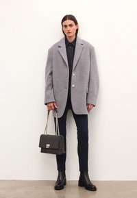 Grijze oversized wollen mix jas met grote revers, twee knopen, gecombineerd met donkere broeken en een gevoerd zwarte handtas met gouden accenten.