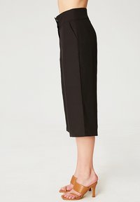 Pantalons noirs raccourcis à taille haute, avec un bouton avant, un design épuré et un tissu lisse. Portés avec des sandales à talons marron.