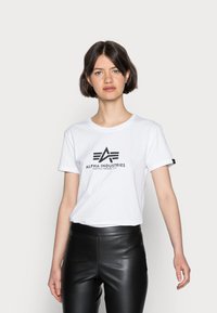 Baltas marškinėlis su trumpomis rankovėmis, su juodu logotipo dizainu "ALPHA INDUSTRIES" ir mažai etikete ant rankovės.