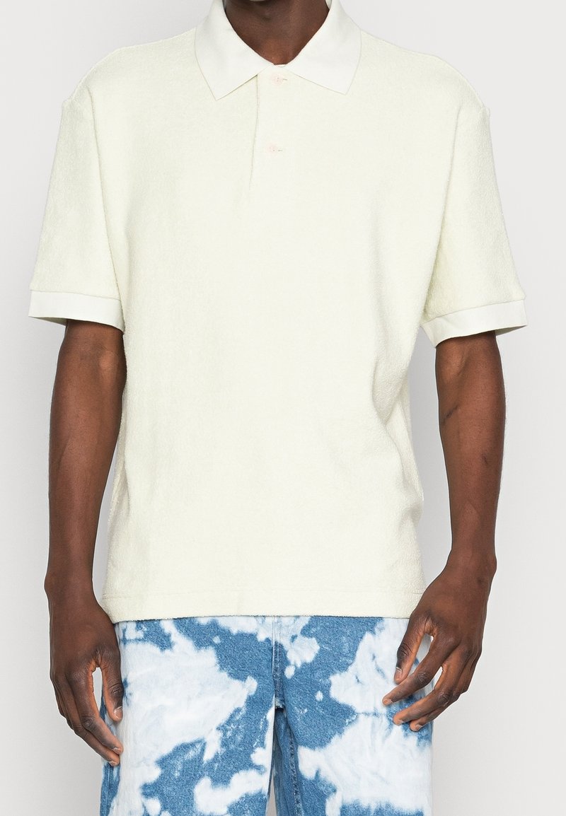 Texturované světle žluté polo tričko s límečkem, krátkými rukávy a dvěma knoflíky, v kombinaci s modrými džínami s tie-dye vzorem.