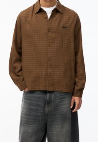 Camisa de cuadros marrón con botones y corte holgado, con un ligero brillo y un pequeño logo en el bolsillo izquierdo del pecho. Combinada con vaqueros gris oscuro.