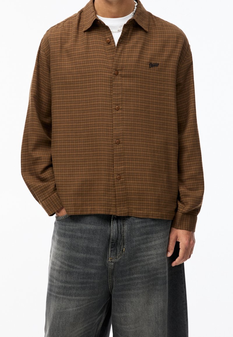 Camisa de cuadros marrón con botones y corte holgado, con un ligero brillo y un pequeño logo en el bolsillo izquierdo del pecho. Combinada con vaqueros gris oscuro.