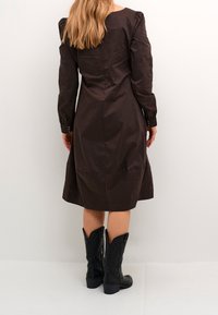 Ruskea, pitkähihainen mekko, jossa on tiukka yläosa ja levenevä hame, nappikiinnityksellä varustetut hihansuut, yhdistetty mustiin, brodeerattuihin cowboy-kengiin.