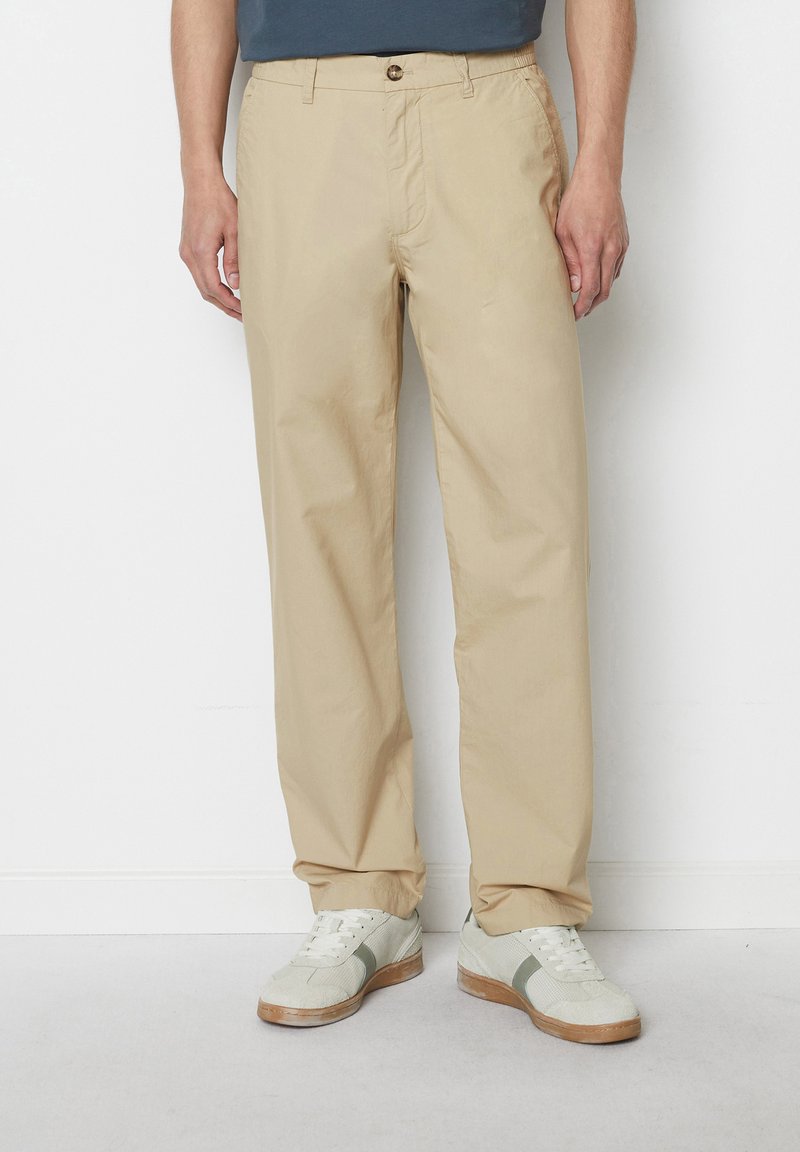 Beige chinos made of lightweight fabric, featuring a straight leg, front pleats, and a single button closure. Paired with textured sneakers.