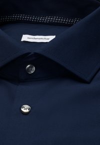 Close-up of a navy blue collared shirt with white buttons, patterned inner collar, and a seidensticker brand label on the inside.