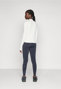 Kvinne med flettet hår iført en hvit langermet topp, mørkegrå leggings og svarte joggesko som står mot en enkel hvit bakgrunn.