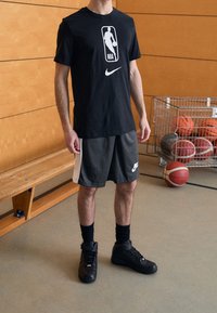 Camiseta negra de la NBA de Nike con logo blanco, pantalones cortos atléticos grises con panel lateral y zapatillas altas negras. Suelo de gimnasio de madera en el fondo.