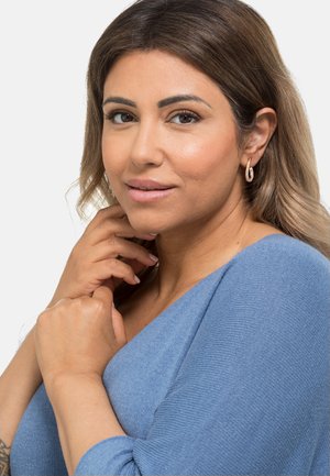 Frau mit langen, gewellten Haaren, die einen hellblauen, gerippten Pullover trägt. Auffällige Details sind die Creolen und die Hand, die auf ihrem Kinn ruht.