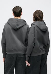 Hanorace gri cu glugă, confecționate dintr-un material moale, cu croială lejeră și manșete cu dungi, purtate de două persoane văzute din spate.