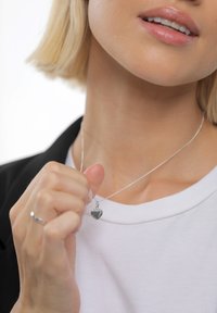 Hõbedast südamekujulise ripatsiga kaelakee peenikesel ahelal, kantud valge särgi kohal. Käsi hoiab kaelakeed, esitledes ripatsi poleeritud pinda.