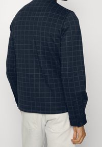 Uomo che indossa una camicia a maniche lunghe a quadri blu scuro, abbinata a pantaloni beige chiaro, mostrato di spalle su uno sfondo semplice.