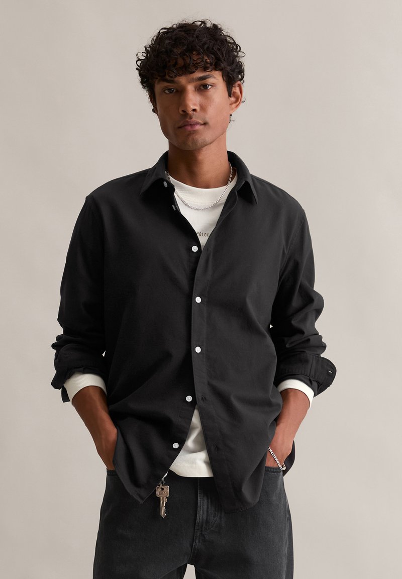 Črna srajca z gumbki in razprtimi ovratniki, prekrita z belo srajco s krojnim ovratnikom, nosena z temnimi kavbojskimi hlačami.