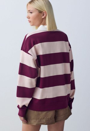 Blonde Person, die nach links blickt, trägt ein locker sitzendes Rugbyshirt mit langen Ärmeln und breiten horizontalen Streifen in Burgund und Hellrosa sowie khakifarbene Shorts.