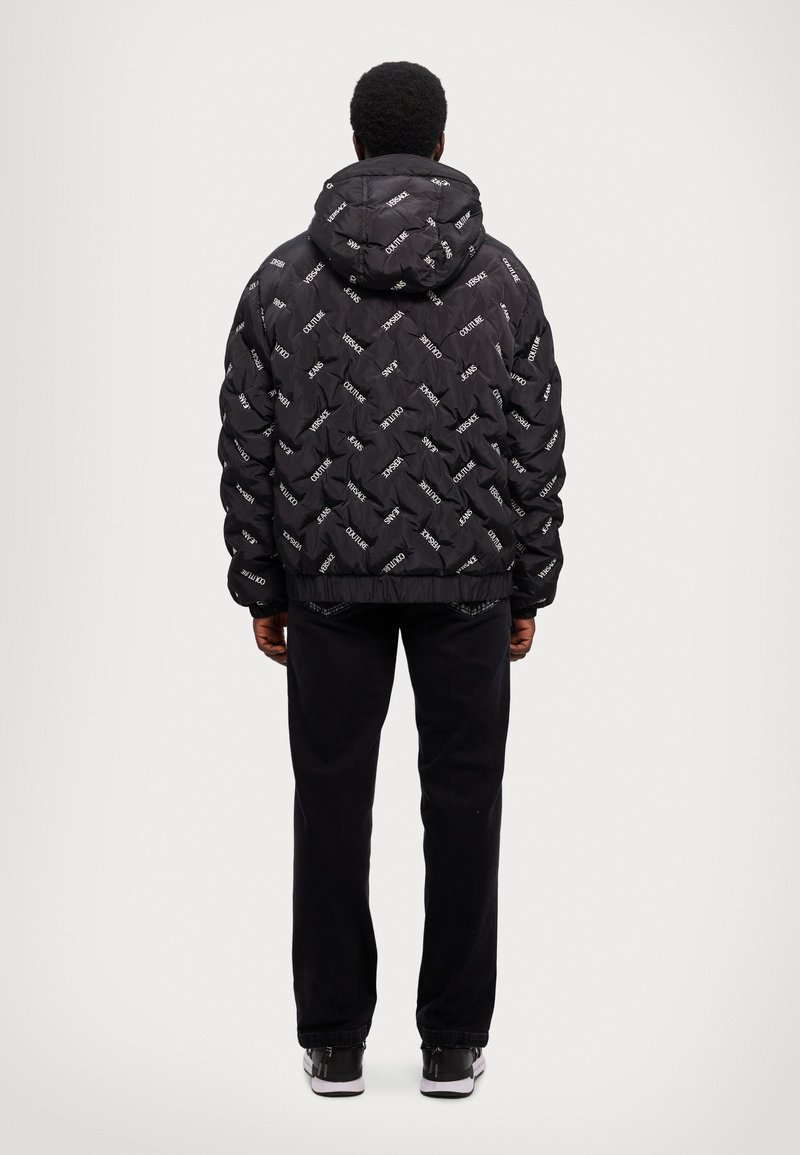 Chaqueta de plumas negra con capucha, que presenta un diseño acolchado y texto del logo en blanco repetido. Combinada con pantalones negros y zapatillas.