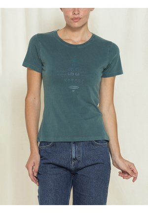 Donna che indossa una maglietta aderente turchese con grafica e jeans blu, in piedi davanti a uno sfondo chiaro e neutro.
