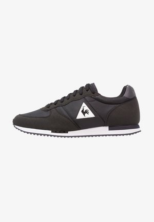 Baskets basses noires avec semelle blanche et logo triangulaire représentant un coq sur le côté, lacées avec des lacets noirs.