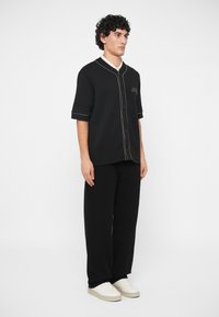 Νεαρός άνδρας με σγουρά μαλλιά, φορώντας μαύρο κοντομάνικο πουκάμισο με άσπρες λεπτομέρειες, μαύρους φαρδύς παντελόνες και άσπρα αθλητικά παπούτσια, στέκεται πλάγια.