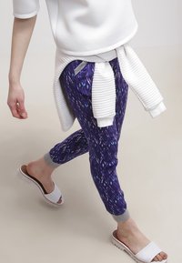 Lila joggingbyxor med ett diamantsmönster, grå manschetter, ihop med en vit sweatshirt och vita slide-sandaler.