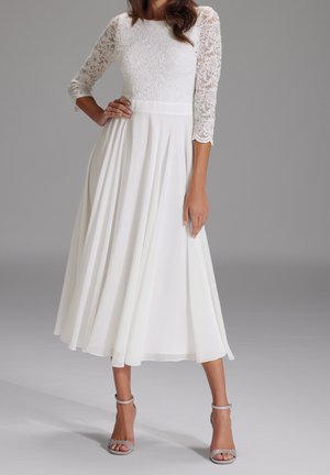 Kobieta ubrana w białą midi sukienkę z koronkowymi rękawami o długości 3/4 i plisowanym dołem, w srebrnych szpilkach na kostkę, stojąca na szarej podłodze.