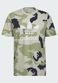 T-shirt verde mimetico con maniche corte, caratterizzato da un logo Adidas bianco sul petto e accenti scuri nel motivo. Materiale in cotone.