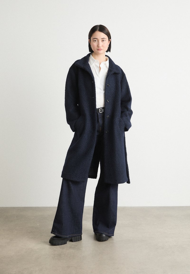 Marineblå oversized frakk laget av teksturert stoff, med knappelukking, åpen krage og sidelommer, parret med brede, mørke jeans.