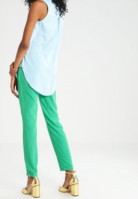 Lys blå ærmeløs top med en længere ryg, parret med grønne tapered bukser og gyldne blokhælesandaler.