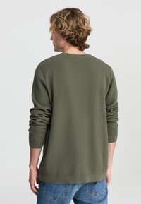 Oliwkowy, textured sweter z długimi rękawami i okrągłym dekoltem. Prosty krój z prążkowanymi mankietami i dołem, noszony z niebieskimi dżinsami.