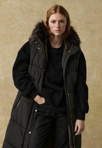 Schwarze Daunenweste mit einer Kapuze aus Kunstpelz, gestepptes Design, kombiniert mit einem schwarzen strukturierten Pullover und schwarzen Hosen. Einfach und praktisch.