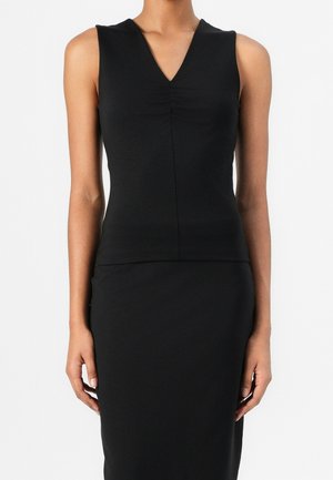 Vrouw draagt een mouwloze zwarte top met V-hals en een bijpassende zwarte rok, staand tegen een effen lichte achtergrond.