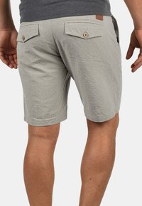 Shorts tissés gris clair avec deux poches arrière et fermetures à bouton, présentant une surface texturée et une longueur mi-thigh.