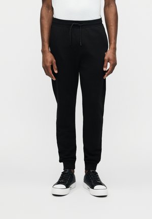 Man met zwarte joggerbroek met trekkoord in de taille en zwarte lage sneakers met witte zolen, staand tegen een witte achtergrond.