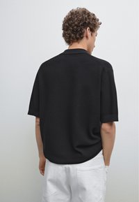 Maglietta polo nera a maniche corte con un design a maglia testurizzata, caratterizzata da una vestibilità rilassata e polsini ripiegati, vista da dietro su uno sfondo chiaro.