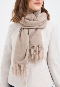 Béžový šál z mäkkej vlny s hustou textúrou a strapcami na okrajoch. Nosený prehodený okolo krku, dopĺňa svetlosivú bundu.