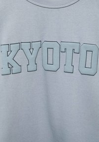 Tessuto azzurro chiaro con grandi lettere in grassetto che scrivono "KYOTO" in un carattere a blocchi nella zona del petto.