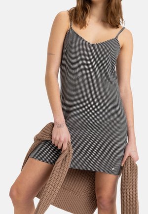 Robe à carreaux noir et blanc avec des fines bretelles, un décolleté droit et une coupe ajustée. Un pull en tricot beige est tenu dans une main.
