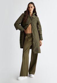 Olijfgroen buitenkleding met een capuchon, gelaagd ontwerp, knopen en ritsdetails, gecombineerd met bijpassende wijde broek en witte sneakers.