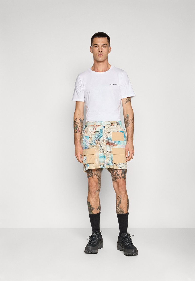Kurzärmliges weißes T-Shirt kombiniert mit gemusterten knielangen Shorts in Beige und Blau, mit Cargo-Taschen und einem Gürtel. Schwarze Socken und Schuhe.