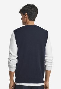 Marinblå ärmlös tröja i mjukt stickat material med rundad halsringning. Bärs över en långärmad vit skjorta som visar en jämn yta.