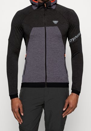 Persoon met een aangesloten, tweekleurige zwart-grijze hoodie met rits, oranje accenten en de tekst "DYNA" op de linkermouw.
