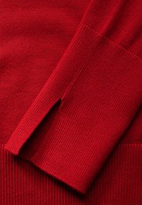 Roter Strickpullover aus Stoff mit gerippten Bündchen und seitlichen Schlitzen. Glatte Textur und gleichmäßige Nähte über das gesamte Material.