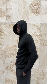Man with hands in pockets wearing black knit hoodie and pants, standing sideways against beige marble tiled wall.