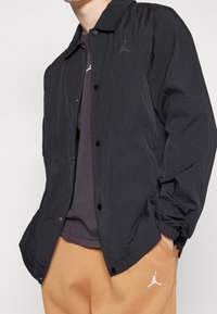 Chaqueta negra de nailon con un diseño estructurado, botones de presión en el frente y un acabado texturizado. Presenta un logo sutil en el lado izquierdo del pecho.