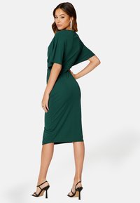 Groene, knie-lange jurk van zachte stof met een gedrapeerde achterkant en korte mouwen. Draagt met zwarte sandalen met bandjes. Gladde textuur zonder patronen.